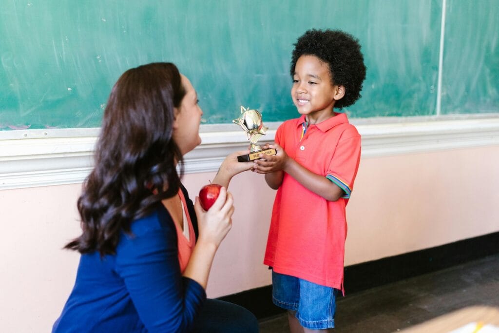 Young student giving a trophy to his teacher, who kneels with a smile and an apple in hand—capturing a heartfelt moment of teacher appreciation.