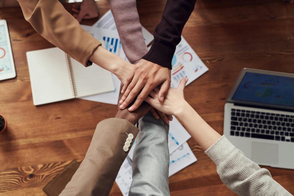 Multiple hands stacked together over a table with papers and a laptop, symbolizing collaboration and shared responsibility in school leadership.